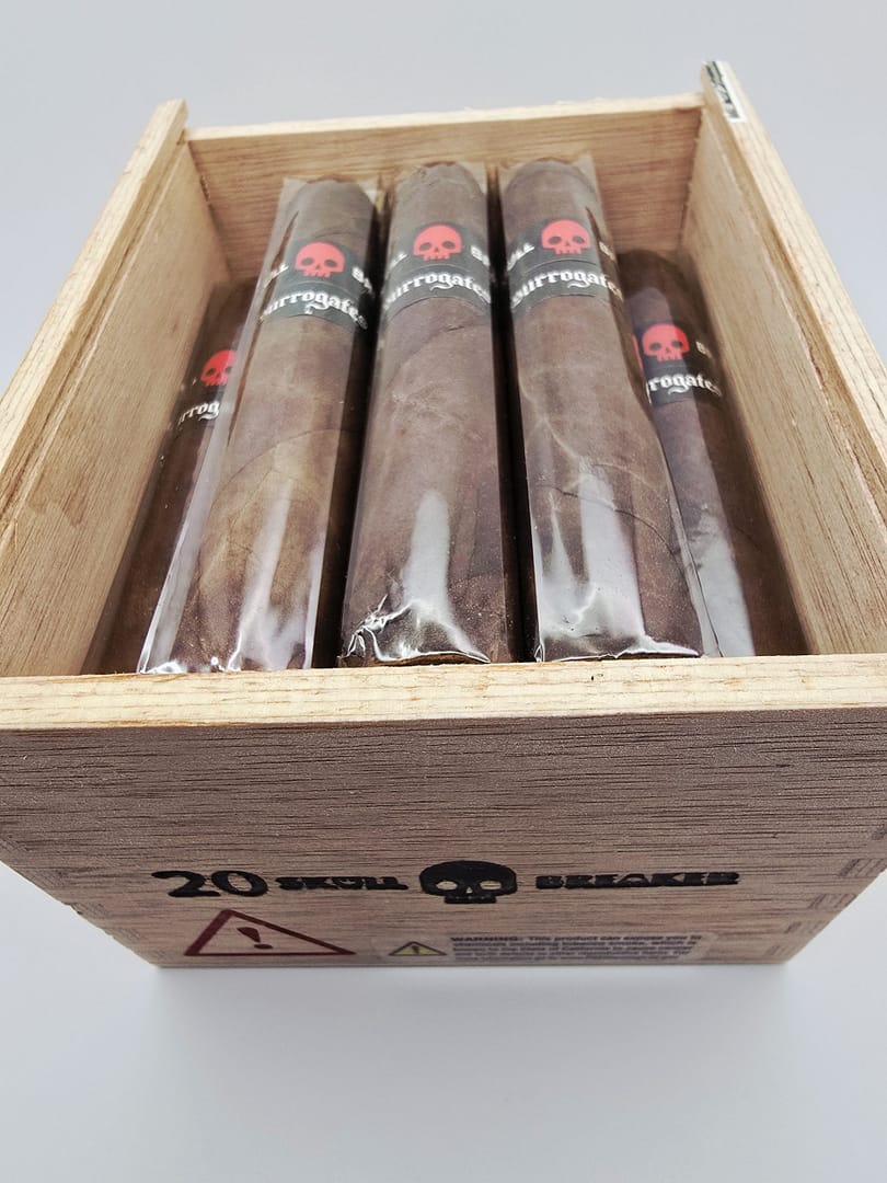 Open box of Tatuaje Surrogates cigars with dark wrappers and bold modern bands arranged in a rustic wooden box.