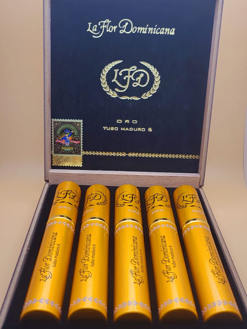 Open box of La Flor Dominicana Oro Tubo Maduro cigars, each housed in a sleek, shiny golden tube, arranged neatly inside a luxurious wooden box.