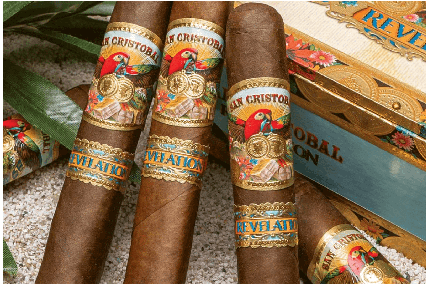 Three San Cristobal Revelation cigars with medium-brown wrappers and ornate bands resting diagonally on a closed wooden cigar box."